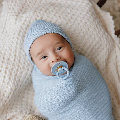 Newborn baby wrapped in light blue swaddle with matching hat, lying on a soft beige blanket using a Luladream premium pacifier. 