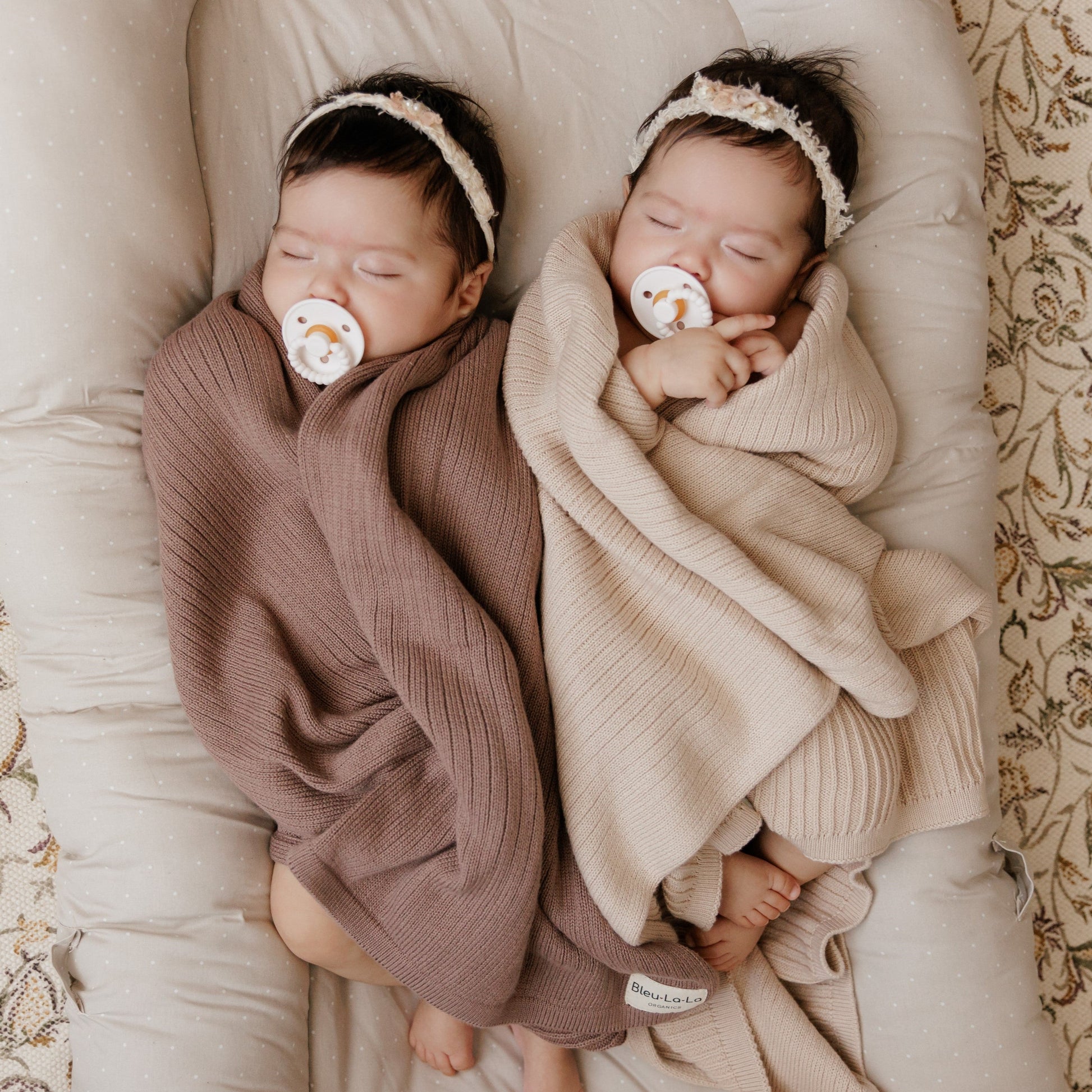 Two babies wrapped in brown and beige blankets with Luladream pacifiers, lying on a soft surface.