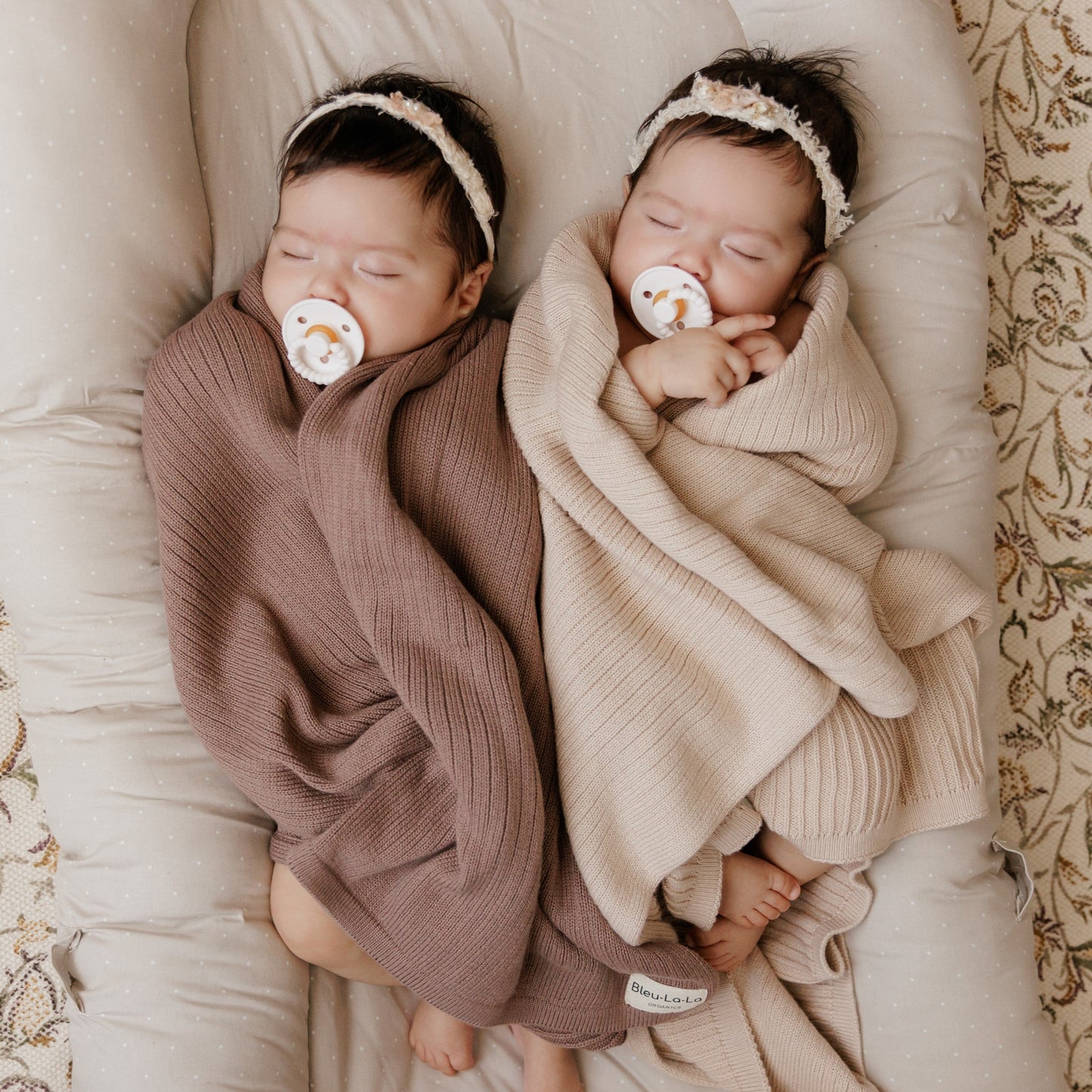 Two babies wrapped in brown and beige blankets with pacifiers, lying on a soft surface.
