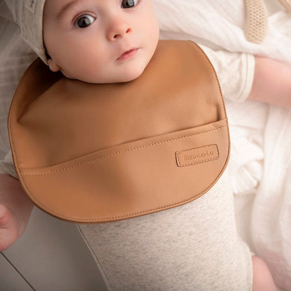 Baby wearing a brown vegan leather easy clean baby bib with a brand label, lying on a soft surface.