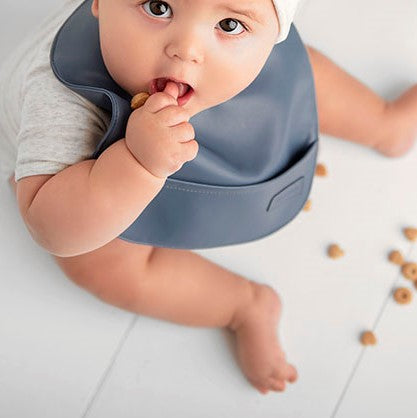 Baby wearing a blue vegan leather easy clean baby bib eating cereal on a white surface