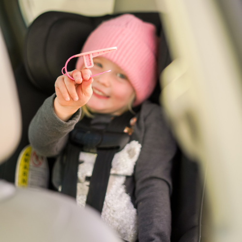 Child in a car seat holding a pink UnBuckleMe safety tool, wearing a pink hat.