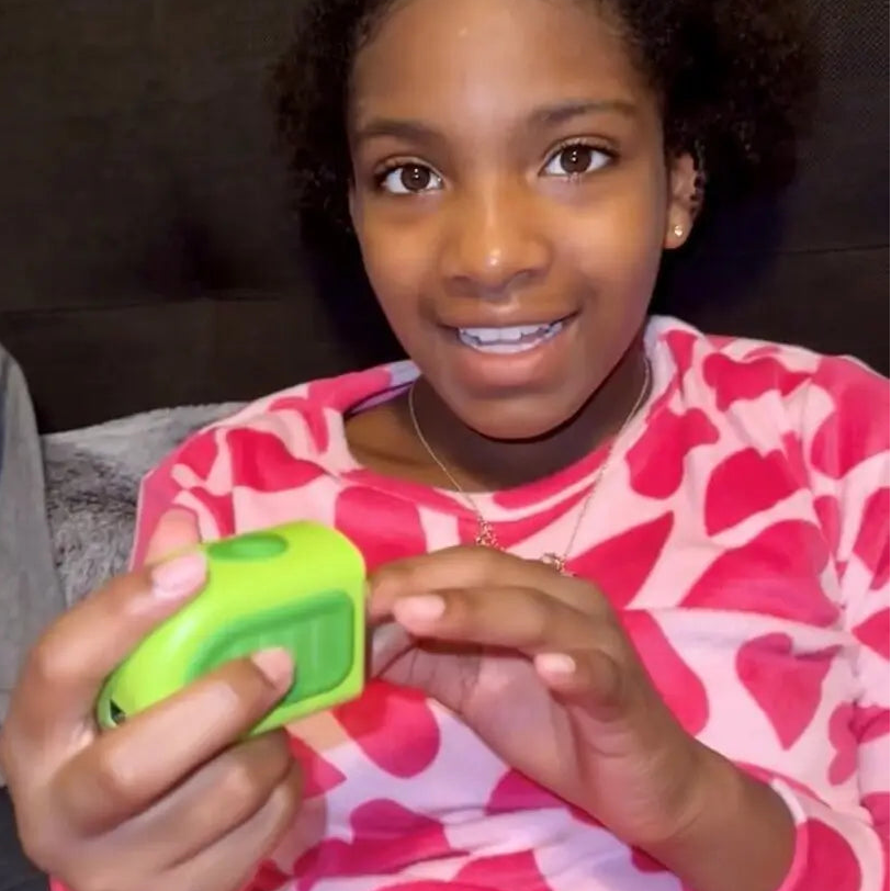 A young girl with curly black hair smiling while holding a Lil Nipper clipping her nails, wearing a pink and white patterned long-sleeve shirt against a dark couch background.
