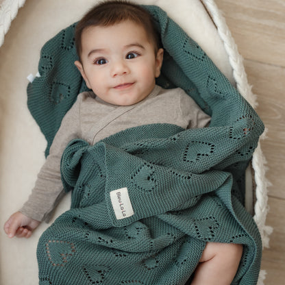 Baby wrapped in a green knitted Bleu-la-la blanket with a visible brand label, sitting in a white crib.