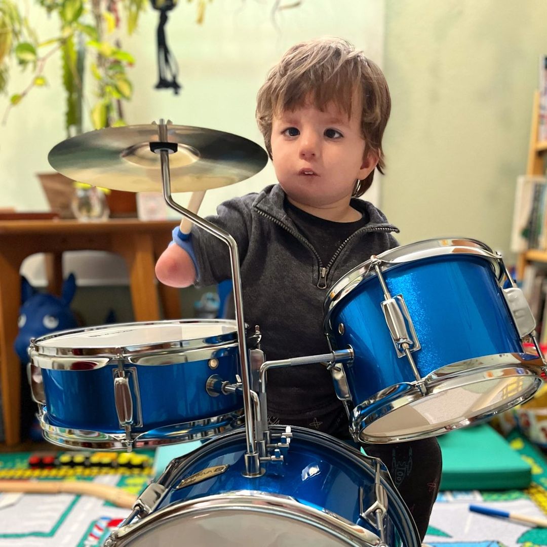 Child with one hand using EaZyHold grippy tool holding drumstick, playing a blue drum set in a room with toys and furniture.