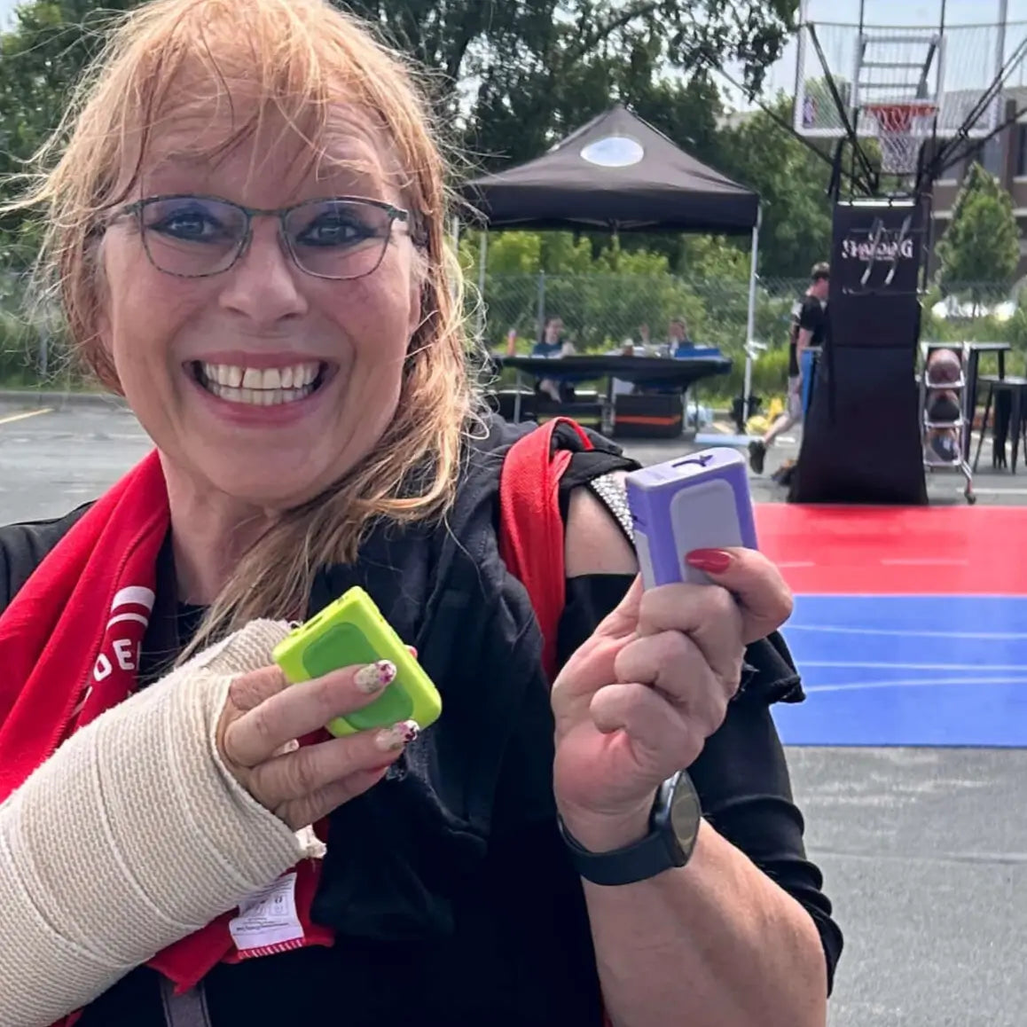 Woman holding a green and a purple Lil Nipper electric nail device on an outdoor basketball court. She's smiling and has an injured hand that's wrapped in bandage.