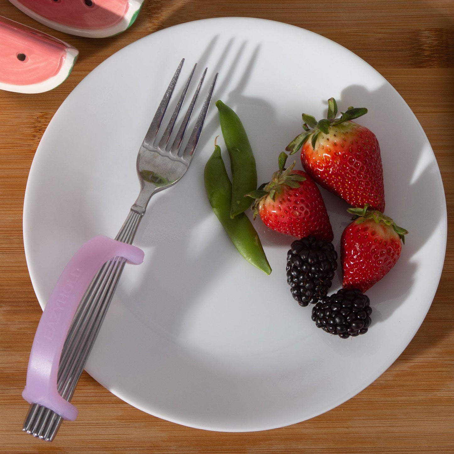 White plate with strawberries, blackberries, green beans, and a fork on a wooden surface with grippy tools on fork.