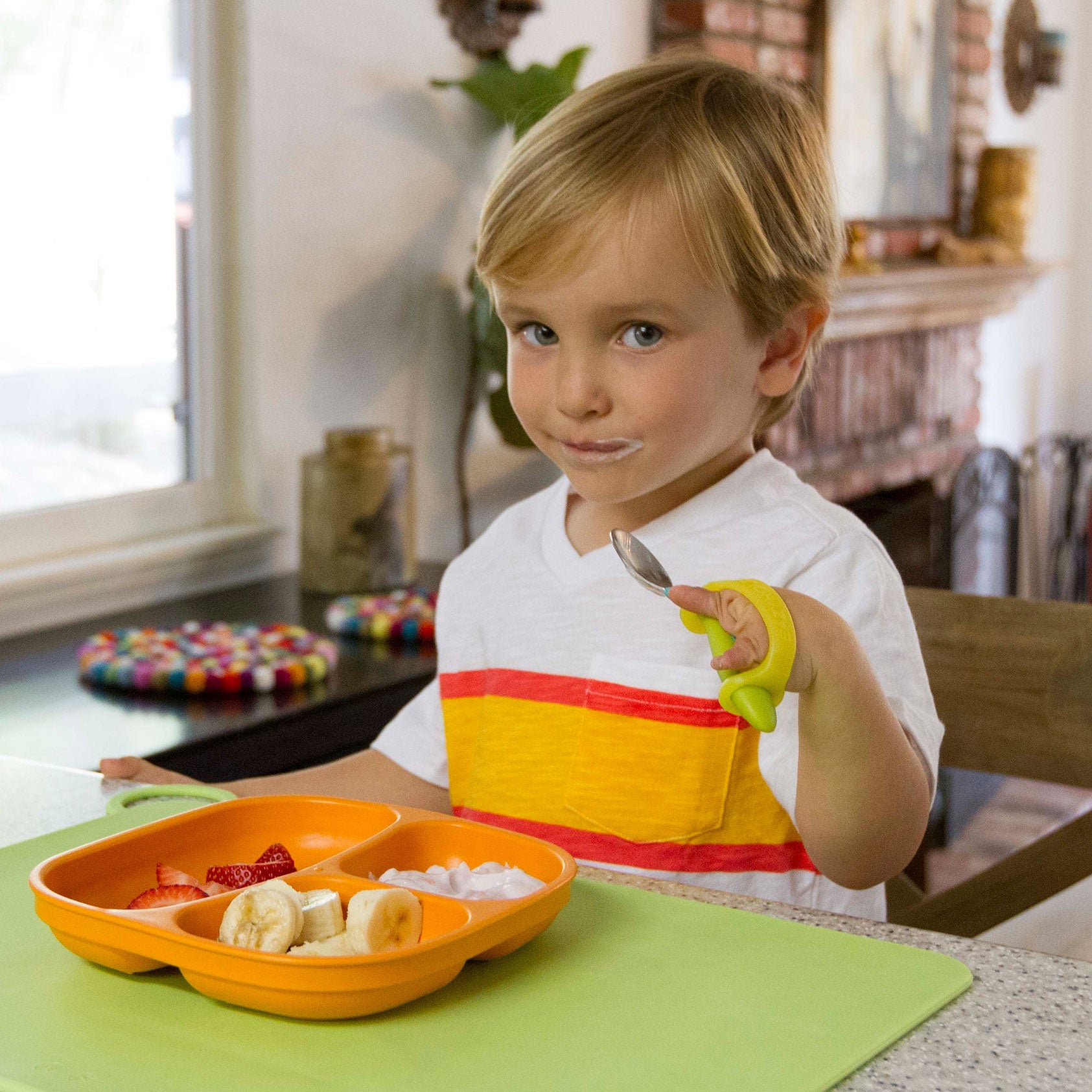 Child sitting at a table with a colorful plate of food and a fork, in a home setting holding a spoon with EaZyHold grippy tool.