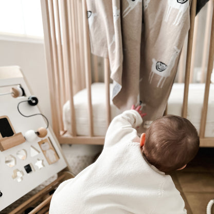 A baby in a white outfit is reaching towards a wooden toy with llama shapes.
